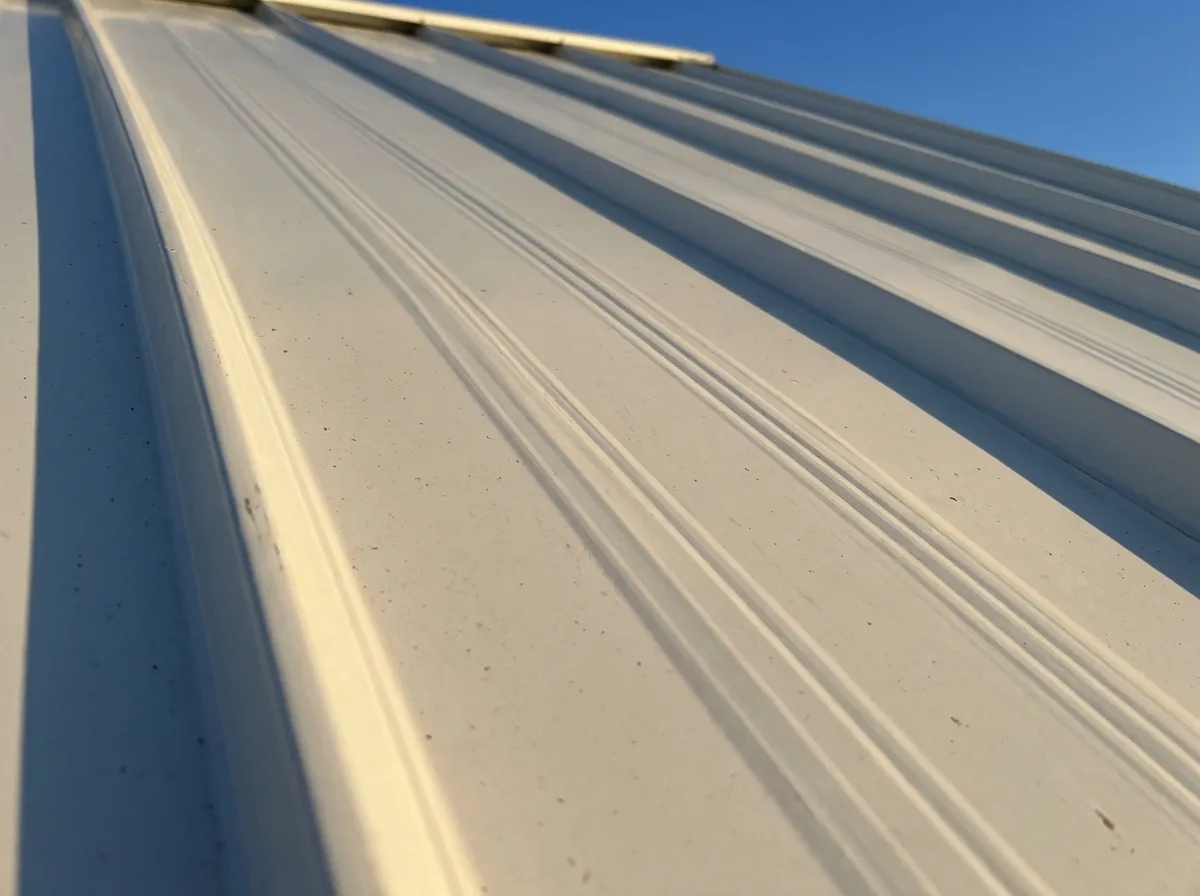 Close view of a standing seam panel with fine pencil ribs pressed lengthwise into its flat surface, catching low sunlight